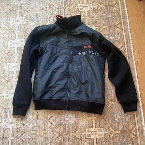 Svart vindjacka från Hugo Boss L - Svart vindjacka från Hugo Boss med dragkedja framtill och hög krage. Jackan har snygga detaljer med BOSS-logga i rött och HUGO BOSS i vitt på bröstet. Materialmix med glansig framsida och mjuka ärmar. Perfekt för en stilren och sportig look.