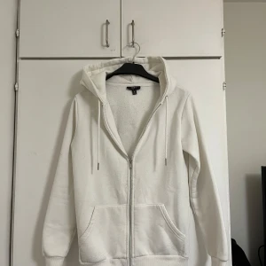 Vit hoodie med dragkedja  XS - Säljer en stilren vit hoodie i storlek XS. Slim fit-modell med dragkedja, huva med snörning och två stora fickor framtill. Mjuk insida och klassisk look som passar perfekt till jeans eller joggers.