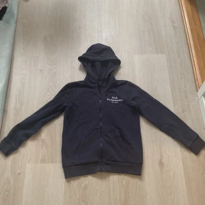 Mörk blå Peak Performance hoodie med dragkedja - Mörk blå  hoodie från Peak Performance med vit logga på bröstet. Tröjan har huva, hel dragkedja och två fickor framtill. Mjuk bomullskänsla och ribbade muddar vid ärmslut och nederkant. Perfekt för chill eller skola.