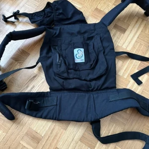 Svart bärsele från Solomom Baby - Säljer en svart bärsele från Solomom Baby med breda axelremmar, justerbart midjebälte och praktisk ficka med dragkedja framtill. Selen är tillverkad i slitstark polyester och har en enkel, stilren design som passar till vardagslivet.