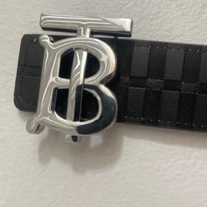 Burberry bälte med rutig design - Snyggt Burberry bälte med stor silvrig TB-logga som spänne. Ena sidan är svart med diskret rutig struktur och präglad riddare, andra sidan har klassiskt beige, svart och röd rutig Burberry-mönster. Bältet är gjort i skinn och har exklusiv känsla.