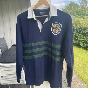 Långärmad pikétröja  Polo Ralph Lauren - Snygg mörkblå långärmad pikétröja från Polo Ralph Lauren med vit krage, gröna ränder över bröstet och ett broderat klubbmärke.  Inga fläckar eller skador. Mindre i storlek. säljer pga för liten storlek. 