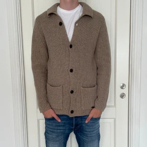 Stickad Cardigan  - Säljer en beige stickad kofta med grov ribbad struktur och svarta knappar framtill. Koftan har två praktiska fickor och en klassisk krage. Perfekt för lager-på-lager och chill stil under kyligare dagar.