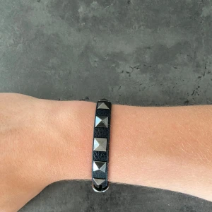 Valentino liknande armband  - Snyggt svart armband i skinnimitation med silvriga pyramidformade nitar längs hela bandet. Justerbar storlek med spänne. Perfekt accessoar för en edgy look och passar till både streetwear och rockiga outfits. Och grish