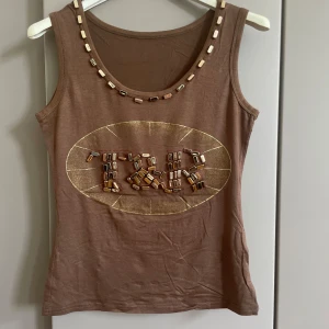 Brun linnetopp med pärldetaljer - Cool brun linnetopp med glansig gulddekor och pärlor både runt halsringningen och på bröstet där det står 'ICB'. Perfekt för dig som gillar unika detaljer och vill sticka ut lite extra. Mjuk och skön bomullskänsla, ärmlös och med rund hals.
