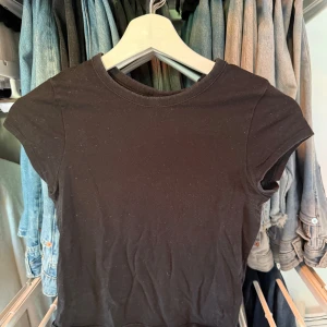 Svart kortärmad topp från Divided XS - En svart, enkel och tight topp från Divided i storlek XS. Modellen är klassisk med rund halsringning och korta ärmar. Perfekt att matcha med jeans eller kjol för en clean look. Materialet är mjukt och stretchigt, vilket gör den riktigt skön att bära.