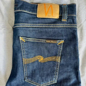 Mörkblå jeans från Nudie Jeans - Snygga mörkblå jeans från Nudie Jeans med klassisk orange söm och logga på bakfickan. Jeansen har raka ben och normal passform, tillverkade i slitstark bomull. Perfekta för dig som gillar stilrena och tidlösa jeans. W33 L30