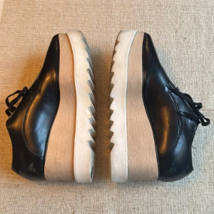 Stella McCartney svarta platåsneakers - Chunky platåsneakers från Stella McCartney i svart syntet med snörning. Skorna har en hög, vågformad plattformsula i trälook och vit, räfflad undersida. Tåformen är spetsig och designen är edgy och modern – perfekta för dig som vill sticka ut.