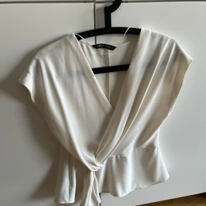 Vit omlottopp från Zara 💕 - Supersnygg vit omlottopp från Zara i storlek S. Toppen har korta ärmar, v-ringning och knyt i midjan som ger en schysst siluett. Tillverkad i ett mjukt, lätt material som faller snyggt. Perfekt när du vill ha en clean och stilren look. Den är i bra skick men den har en liten fläck på ryggen (finns bild i annonsen). 💕