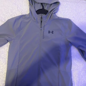 Grå vindjacka från Under Armour - Snygg grå vindjacka från Under Armour med huva och hel dragkedja framtill. Jackan har diskret logga på bröstet och är tillverkad i ett lätt, syntetiskt material som passar perfekt för träning eller när du vill ha en sportig look. Långärmad och med praktiska fickor på sidorna. ‼️storlek-S‼️Använts 2-3 ggr. Liten fläck längst ner i vänstra armen! Obs säljs bara i Jkpg