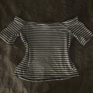 Randig offshoulder topp i bomull - Snygg vit och svart randig topp med offshoulder-modell och korta ärmar. Figurnära passform som framhäver siluetten, perfekt för en trendig och avslappnad look. Gjord i mjuk bomull som känns skön mot huden.