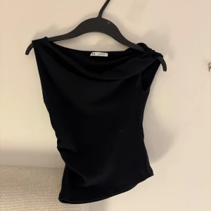 Svart ärmlös topp från Zara - Svart, stilren ärmlös topp från Zara med snygg draperad halsringning. Toppen har en tight passform och är tillverkad i ett mjukt, stretchigt material som sitter skönt mot kroppen. Perfekt att matcha med jeans eller kjol för en trendig look.
