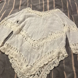 Vit boho blus med fransdetaljer - Superfin vit blus i tunn bomull med boho-vibbar. Blusen har långa ärmar och är dekorerad med virkade fransar i zigzag-mönster längs ärmar, halsringning och nederkant. Perfekt för sommarens festivaler eller strandhäng.