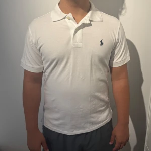 Vit pikétröja från Polo Ralph Lauren - Klassisk vit pikétröja från Polo Ralph Lauren med blå broderad logga på bröstet. Tröjan har korta ärmar, krage och knäppning med två knappar. Tillverkad i bomull och har en slim passform som sitter snyggt på kroppen.