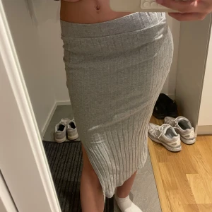 Grå ribbad långkjol med slits - Säljer en grå ribbad långkjol med slits på sidan. Kjolen är figurnära och har en bekväm midjeresår. Perfekt för dig som gillar en enkel men snygg look. Materialet är mjukt och stretchigt, vilket gör den superbekväm att bära.