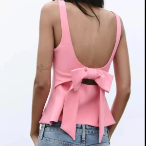 Rosa topp med rosett från Zara - Supersöt rosa topp från Zara med fyrkantig ringning och breda axelband. Toppen har öppen rygg och en stor rosett bak som ger en extra snygg detalj. Figurnära passform och lätt utsvängd nertill. Perfekt för dig som vill sticka ut lite extra!