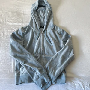 Ljusblå velour hoodie Juicy Couture - Supermjuk ljusblå hoodie från Juicy Couture i velour med dragkedja framtill och snörning i huvan. Klassisk logga broderad på bröstet och ribbade muddar. Perfekt för en chill och trendig look.
