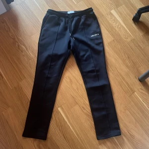 Svarta mjukisbyxor från Les Deux - Svarta mjukisbyxor från Les Deux med raka ben och snygga pressveck. Byxorna har dragsko i midjan och diskret logga på ena benet. Sköna och stilrena, perfekta för en chill och clean look.