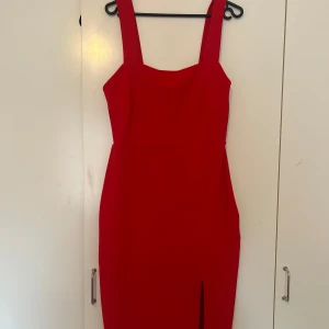 Röd figurnära långklänning - Snygg röd långklänning med breda axelband och en tight passform. Klänningen har en stilren design med slits framtill som ger en extra edge. Perfekt för dig som vill sticka ut med en klassisk men ändå modern look.