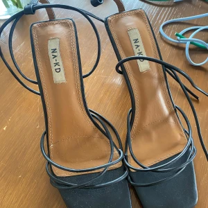 Svarta klackar från NA-KD - Svarta klackar från NA-KD med fyrkantig tå, tunna remmar som snöras runt benet och hög klack. Snygg design med bruna innersulor och lackad finish på klacken. Perfekta för dig som gillar trendiga statement-skor. Skorna har slitning som ni ser på bilderna 