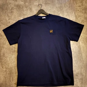 Mörkblå Loewe t-shirt med broderi - Snygg mörkblå t-shirt från Loewe i bomull med klassisk passform. På bröstet finns ett broderat Loewe-logo i svart och gult som ger en clean men ändå unik vibe. Perfekt till jeans eller shorts för en stilren look.. passar även L jag är 190 och passar mig perfekt också, den är oversized