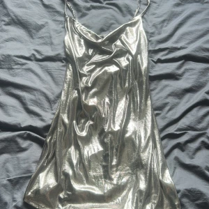 Glittrig silver kortklänning Nowomenslabel - Supersnygg glittrig silver kortklänning från Nowomenslabel med tunna axelband och draperad urringning. Perfekt för dig som vill sticka ut med en metallic look. Materialet är glansigt och följsamt, vilket ger en cool och trendig vibe.