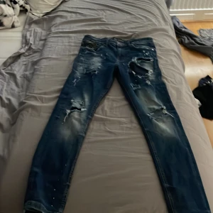 Blå ripped jeans Supply & Demand - Säljer ett par blå jeans från Supply & Demand med slitna detaljer och hål på benen för en riktigt edgy look. Jeansen har normal passform och klassisk femficksdesign. Perfekta för dig som gillar streetstyle och vill sticka ut lite extra.