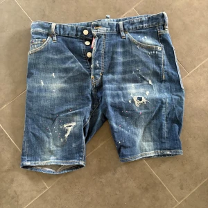 Blå jeansshorts med slitningar - Snygga blå jeansshorts från Dsquared2 med slitna detaljer, färgstänk och patch baktill. Klassisk femficksmodell med knappar i gylfen och coola slitningar framtill och baktill. Perfekta för dig som gillar streetstyle och vill sticka ut lite extra.