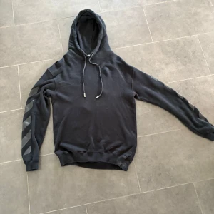 Svart Off-White hoodie med tryck - Svart hoodie från Off-White med ikoniska pilar och ränder i grått på ärmarna och stor logga på ryggen. Tröjan har huva med snörning och ribbade muddar. Perfekt för dig som gillar streetwear och vill sticka ut med en stilren look.