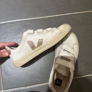 Vita Veja sneakers  - Snygga vita sneakers från Veja med guldiga detaljer på häl och sidor. Skorna har tre kardborreband för enkel påtagning och en klassisk rund tå. Tillverkade i skinn med platt sula, perfekta för en clean och trendig look.