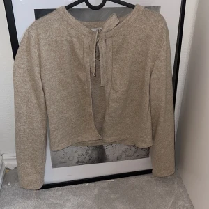 Beige kofta med knyt från 157 - Säljer en beige kofta från 157 i mjukt material. Koftan har lång ärm och knytning framtill vid halsen för en chill och trendig look. Perfekt att slänga över en topp när det är lite kyligt ute.