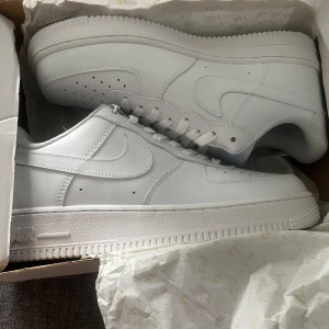 Nike Air Force 1 helvita sneakers - Klassiska Nike Air Force 1 sneakers i helvitt skinn med perforerad tå, rund tå och platt sula. Snygg och clean look med vita snören och diskret AF1-detalj i metall framtill. Perfekta för dig som gillar tidlös streetstyle.