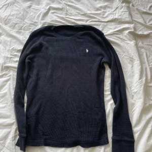 Mörkblå långärmad tröja Polo Ralph Lauren - Mörkblå långärmad tröja från Polo Ralph Lauren i storlek XL. Tröjan har ett diskret rutmönster i strukturen och en liten broderad logga på bröstet. Tillverkad i mjuk bomull och har ribbade muddar vid ärmslut och hals.