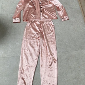 Rosa satinpyjamas från SHEIN - Supermjuk och glansig rosa pyjamas från SHEIN i satinlook. Setet har långärmad skjorta med svarta knappar och piping, samt matchande byxor med resår i midjan. Perfekt för mysiga kvällar hemma.