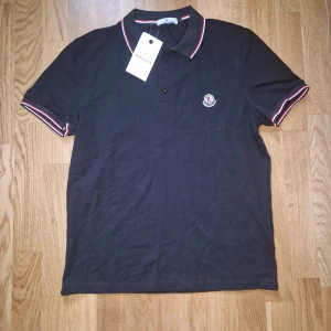 Svart Moncler pikétröja med röd/vita detaljer - Kan passa både M och S. Snygg svart pikétröja från Moncler med klassisk krage och knappar framtill. Rödvita ränder på krage och ärmslut ger en sportig vibe. Broderad Moncler-logga på bröstet. Perfekt för dig som gillar stilrena och exklusiva plagg.