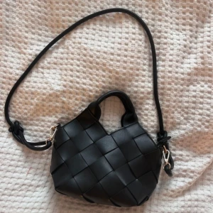 Svart axelväska från Pitaya i skinn - Snygg svart axelväska från Pitaya i flätad skinnimitation. Väskan har både handtag och lång axelrem, samt guldfärgade metalldetaljer och dragkedja upptill. 