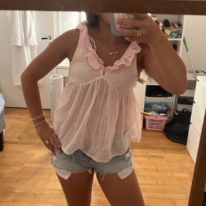 Ljusrosa volangig blus i bomull - Supersöt ljusrosa blus med volangkant vid v-ringningen och luftig, flowy passform. Ärmlös modell i tunt, skrynkligt bomullstyg som känns perfekt för varma dagar. Ger en romantisk och chill vibe till din outfit. Säljer den i storlek 170 men den passar även xs💞