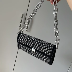 Svart axelväska med silverdetaljer - Snygg svart glittrig axelväska chh en chunky kedja som axelrem. Väskan har rektangulär form, lock med silverspänne och cool partyvibe. Perfekt för dig som vill sticka ut med accessoarer.