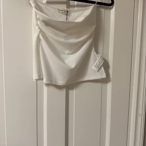 Vit axelbandslös tubtopp Pull&Bear - Snygg vit tubtopp från Pull&Bear med draperad framsida. Axelbandslös och croppad modell som passar perfekt till höga jeans eller kjol. Enkel och stilren design som är lätt att matcha.