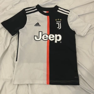 Juventus Ronaldo 7 matchtröja Adidas S - Juventus fotbollströja med Ronaldo 7 på ryggen. Snygg design i svart och vitt med röd detalj i mitten, klassiska Adidas-ränder på axlarna och Jeep-logga framtill. Tillverkad i lätt och ventilerande Climalite-material, perfekt för fotbollsplanen.