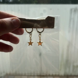 Guldiga örhängen med stjärnhänge - Snygga örhängen i guld med små stjärnhängen som dinglar från en tunn kedja. Perfekta för dig som vill ha en cool och trendig detalj till din look. Stjärnorna ger en lekfull vibe och örhängena är lätta att matcha med andra smycken.