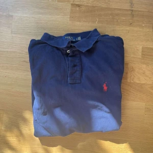 Ralph Lauren piké - Klassisk marinblå pikétröja från Polo Ralph Lauren i bomull. Slim fit-modell med korta ärmar, krage och knäppning framtill. Står XL i size men skulle inte hålla med och säger att det snarare sitter som M-L. Ikoniska röda loggan broderad på bröstet. Perfekt för en clean och stilren look. 
