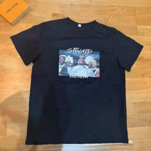 Svart t-shirt med tryck 'STWAID WEALTH' - Svart t-shirt i storlek M med ett stort färgglatt tryck på bröstet som visar personer med sedelbuntar och texten 'STWAID WEALTH' i rosa. Klassisk rund hals och korta ärmar. Perfekt för dig som gillar streetwear och statement-plagg.