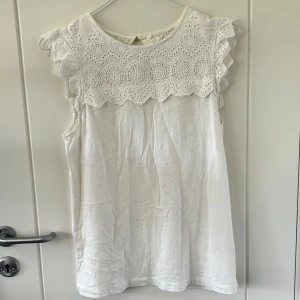 Vit blus från Lindex strl S - Superfin vit blus från Lindex med broderade detaljer och volang vid axlarna🤍Ärmlös modell med rund halsringning och snygga guldknappar i ryggen 🤍Tillverkad i bomull🤍 står ej storlek men gissar på S🤍säljs pga att den är för liten