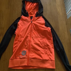WOXO orange och svart vindjacka - Snygg orange och svart vindjacka från WOXO, designad av KappAhl. Jackan har huva, hel dragkedja framtill, två fickor och en extra ficka bak. Tillverkad i polyester med sportig look och tryckt logga både fram och bak. Perfekt för dig som gillar streetstyle.