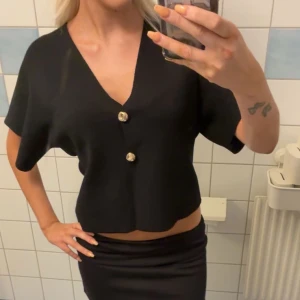 Svart tröja med guldknappar - Svart croppad blus med v-ringning och två stora guldiga knappar framtill. Modellen har korta, vida ärmar 