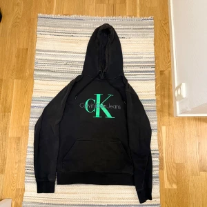 Svart hoodie från Calvin Klein Jeans - Svart hoodie från Calvin Klein Jeans med stor grön CK-logga och text på bröstet. Klassisk känguruficka framtill, snörning i huvan och ribbade muddar. Perfekt för en chill och stilren look. Limiterad, hör av dig vid frågor!