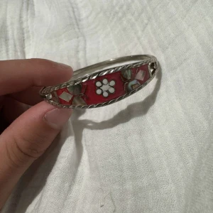 Silvrigt armband med blommönster - Unikt armband i silverfärg med rosa bas och blommönster i vitt och pärlemor. Armbandet har en dekorativ kant och detaljer i olika former, vilket ger en cool och bohemisk vibe. Perfekt för dig som gillar färg och vill sticka ut lite extra.