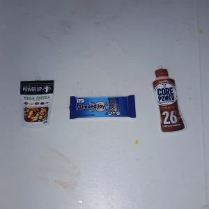 Protein saker berlocker - Bilden visar tre miniatyrförpackningar av snacks och dryck: Power Up Mega Omega, Almond Joy och Core Power Chocolate.