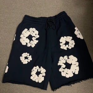 Marin blåa denim tears shorts - Mörkblå shorts från Denim Tears med vita blommiga tryck över hela plagget. Modellen har elastisk midja med snörning och råa kanter nertill för en avslappnad vibe. Perfekta för sommaren och riktigt snygga till sneakers eller sandaler.